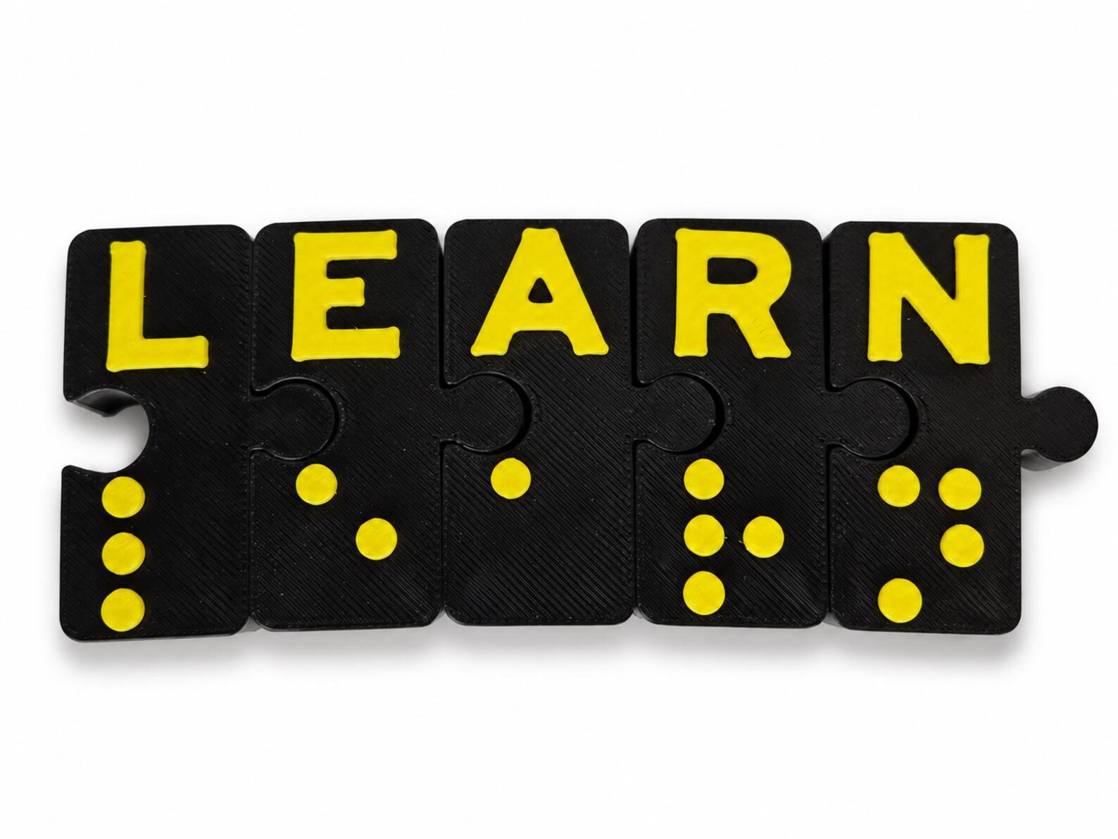 Braille Connex interlocking puzzle blocks spelling LEARN in raised yellow print letters, with the matching yellow Braille dots below each letter.